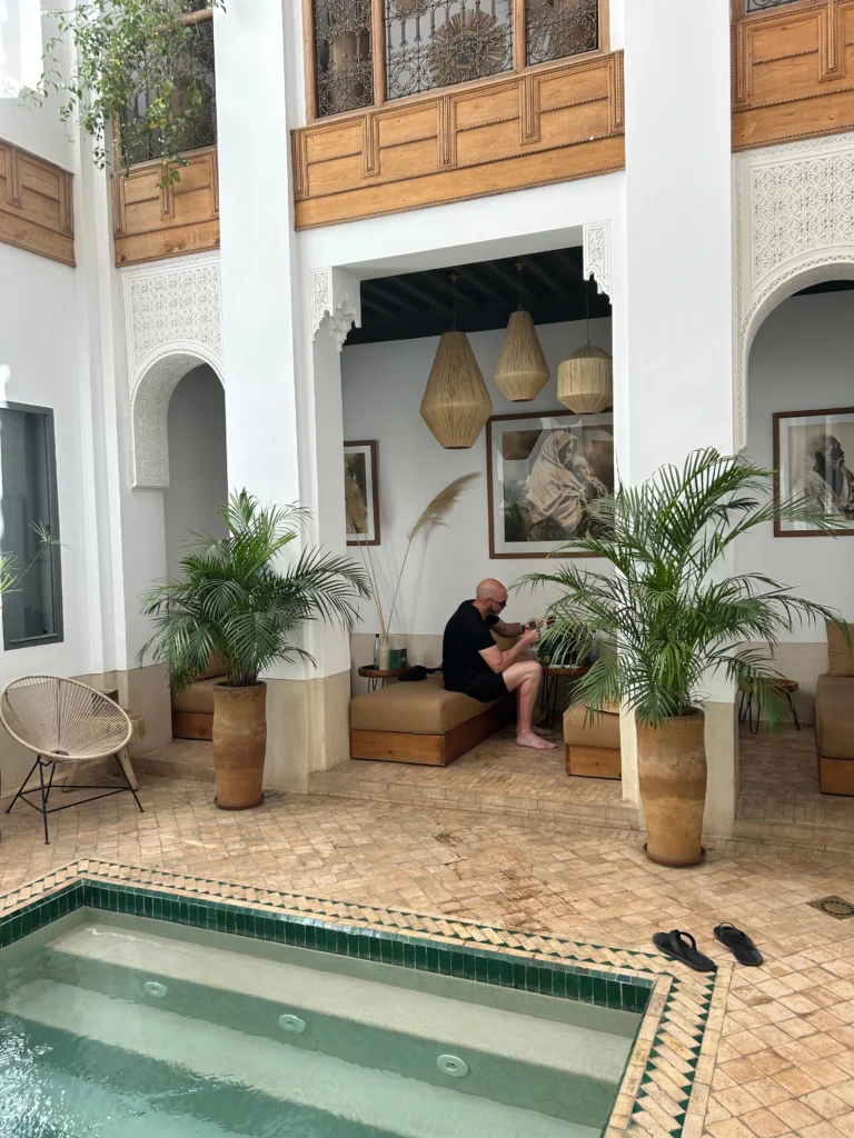 A beautiful luxury riad in Marrakech, showcasing intricate Moorish architecture with a central courtyard filled with lush greenery and vibrant flowers. In the foreground, a serene fountain reflects the intricate tile work and rich colors of the surrounding architecture. The middle ground features elegantly furnished lounges with ornate textiles and traditional Moroccan lanterns casting a warm glow. In the background, glimpses of the bustling Marrakech streets contrast with the tranquility of the riad's interior. Soft, golden hour lighting enhances the ambience, creating an inviting and luxurious atmosphere. The image captures the essence of relaxation and unique cultural heritage, perfect for highlighting the special features of luxury riads.