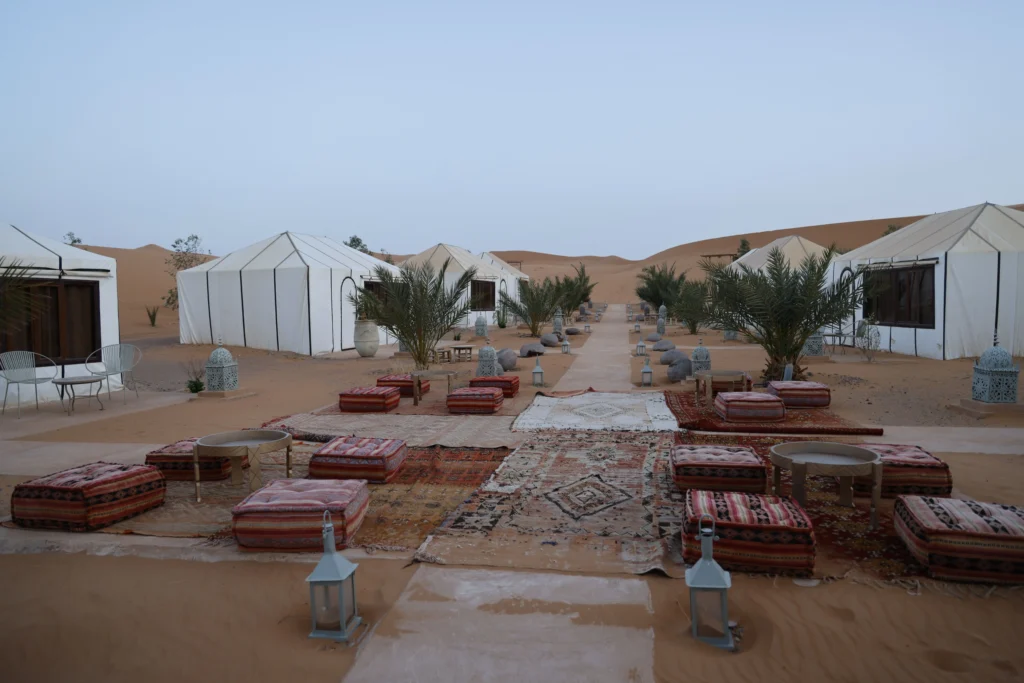 A luxurious desert camp in Morocco, nestled among golden sand dunes at sunset. In the foreground, see elegantly designed tents with intricate patterns and plush furnishings, including vibrant cushions and traditional Moroccan lanterns casting warm, inviting light. Middle ground features a cozy fire pit surrounded by comfortable seating, where guests relax in stylish but casual clothing, enjoying the serene atmosphere. The background showcases rolling sand dunes under a colorful sky transitioning from orange to deep purple. Soft, diffused lighting enhances the serene and inviting mood, with a few palm trees swaying gently in the breeze. Capture this idyllic setting from a slightly elevated angle to emphasize the camp's luxury and the enchanting desert landscape. A luxurious desert camp in Morocco