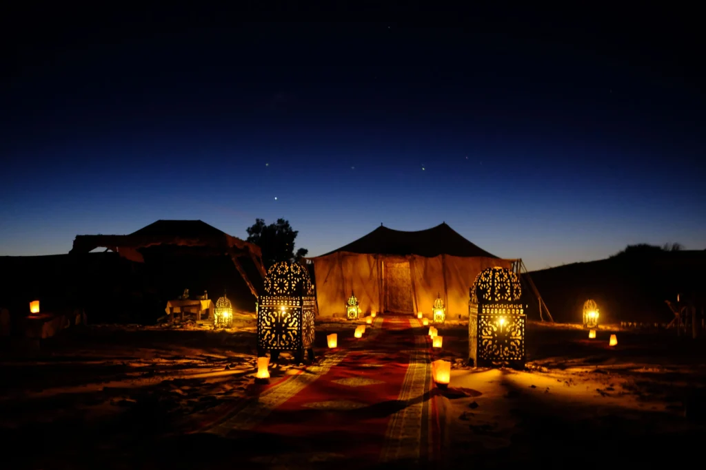 A luxurious desert camp in Morocco set against the vast golden sands of the Sahara. In the foreground, elegantly designed tents with rich, intricate patterns and soft ambient lighting create a warm, inviting atmosphere. Plush cushions and low tables adorned with traditional Moroccan tea sets enhance the cozy feel. In the middle ground, a campfire flickers gently, surrounded by comfortable seating, inviting guests to relax under the starry night sky. The backdrop features towering sand dunes under a breathtaking sunset, with hues of orange and purple blending into a serene twilight. The scene evokes a sense of adventure and tranquility, ideal for an unforgettable stay. The image should capture the essence of opulence amid nature, with soft, diffused lighting to create a magical atmosphere. A luxurious desert camp in Morocco set against the vast golden sands of the Sahara. In the foreground, elegantly designed tents with rich, intricate patterns and soft ambient lighting create a warm, inviting atmosphere. Plush cushions and low tables adorned with traditional Moroccan tea sets enhance the cozy feel. In the middle ground, a campfire flickers gently, surrounded by comfortable seating, inviting guests to relax under the starry night sky. The backdrop features towering sand dunes under a breathtaking sunset, with hues of orange and purple blending into a serene twilight. The scene evokes a sense of adventure and tranquility, ideal for an unforgettable stay. The image should capture the essence of opulence amid nature, with soft, diffused lighting to create a magical atmosphere.