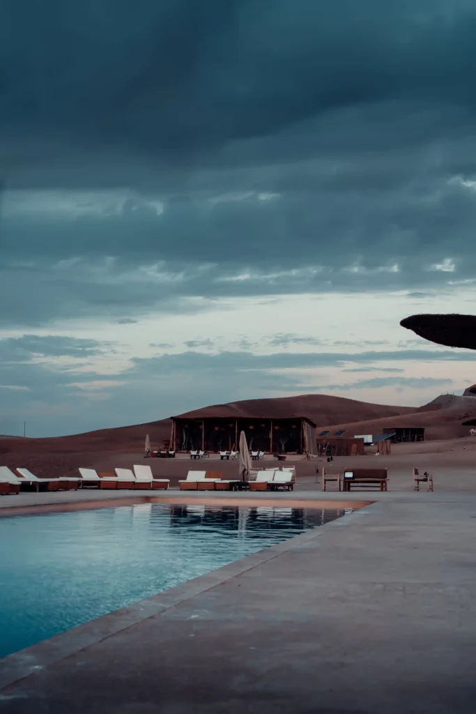 A luxurious desert camp in Morocco at sunset, showcasing elegantly styled tents adorned with rich textiles and intricate lanterns illuminating the scene. In the foreground, a beautifully set dining area with plush cushions and a low table, laden with traditional Moroccan dishes and aromatic teas. The middle ground features gracefully designed tents with flaps open, revealing soft interiors and luxurious bedding. In the background, vast dunes gently lit by the warm sun, creating a serene and magical atmosphere. The sky transitions to vibrant hues of orange and purple, enhancing the enchanting mood. Shot with a soft focus lens to capture the warm glow, conveying an inviting and unforgettable experience in this desert paradise. A luxurious desert camp in Morocco at sunset, showcasing elegantly styled tents adorned with rich textiles and intricate lanterns illuminating the scene. In the foreground, a beautifully set dining area with plush cushions and a low table, laden with traditional Moroccan dishes and aromatic teas. The middle ground features gracefully designed tents with flaps open, revealing soft interiors and luxurious bedding. In the background, vast dunes gently lit by the warm sun, creating a serene and magical atmosphere. The sky transitions to vibrant hues of orange and purple, enhancing the enchanting mood. Shot with a soft focus lens to capture the warm glow, conveying an inviting and unforgettable experience in this desert paradise.