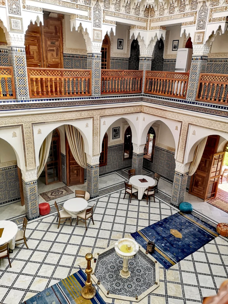 A stunning interior view of a luxurious Moroccan riad in Marrakech, featuring intricately designed tile work, vibrant mosaic patterns, and lush indoor gardens. In the foreground, a richly decorated courtyard with a serene fountain, surrounded by elegant seating areas adorned with plush cushions. The middle section showcases the ornate details of traditional wooden doors, intricate plasterwork, and glowing lanterns casting warm, inviting light. In the background, tall palm trees and blue sky peek through arched windows, enhancing the exotic atmosphere. The scene is bathed in soft, natural light, creating a tranquil and welcoming mood, ideal for highlighting the beauty and elegance of luxury riads in Marrakech.