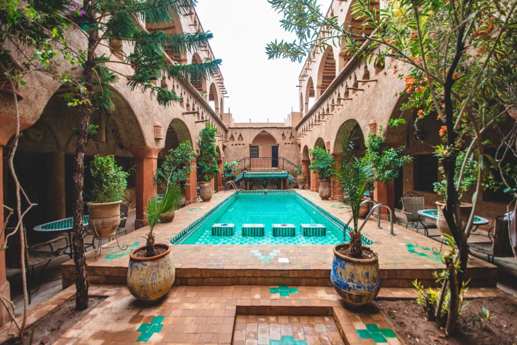 A stunning traditional Moroccan riad, showcasing intricate tile work and lush greenery. In the foreground, an elegant courtyard filled with vibrant flowers and a sparkling fountain. The middle ground features ornate archways and beautifully carved wooden doors, typical of Moroccan architecture. The background offers glimpses of a serene blue sky framed by colorful mosaics. The lighting is warm and inviting, casting soft shadows that enhance the textures of the riad’s walls. The scene conveys a tranquil and luxurious atmosphere, perfect for showcasing the charm of Moroccan hospitality. Capture this image from a low angle to emphasize the stature of the riad, creating a sense of intimacy and beauty.