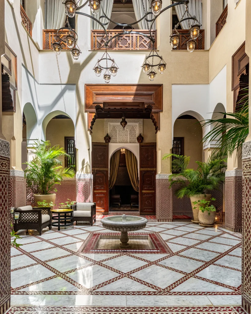 A stunning traditional Moroccan riad, showcasing intricate tile work and lush greenery. In the foreground, an elegant courtyard filled with vibrant flowers and a sparkling fountain. The middle ground features ornate archways and beautifully carved wooden doors, typical of Moroccan architecture. The background offers glimpses of a serene blue sky framed by colorful mosaics. The lighting is warm and inviting, casting soft shadows that enhance the textures of the riad’s walls. The scene conveys a tranquil and luxurious atmosphere, perfect for showcasing the charm of Moroccan hospitality. Capture this image from a low angle to emphasize the stature of the riad, creating a sense of intimacy and beauty.